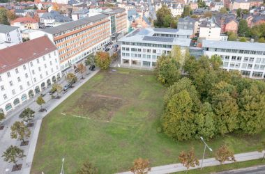Drohnenaufnahme mit Blick auf das Woolworth-Gelände. Eine Fläche mit Wiese und Bäumen. Dahinter sind Häuser und Gebäude der Stadt zu sehen.