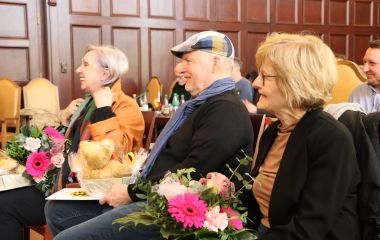 Drei Personen sitzen nebeneinander und halten ein Präsent und Blumen in den Händen.