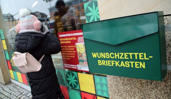 An der Hauswand neben einem Schaufenster hängt ein grüner Briefkasten. Auf dem Briefkasten steht Wunschzettelbriefkasten. Ein Mädchen steht vor dem Schaufenster und sieht hinein.