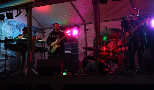 Eine Band tritt auf einer Bühne auf. Es ist dunkel. Bunte Scheinwerfer erhellen das Bild.