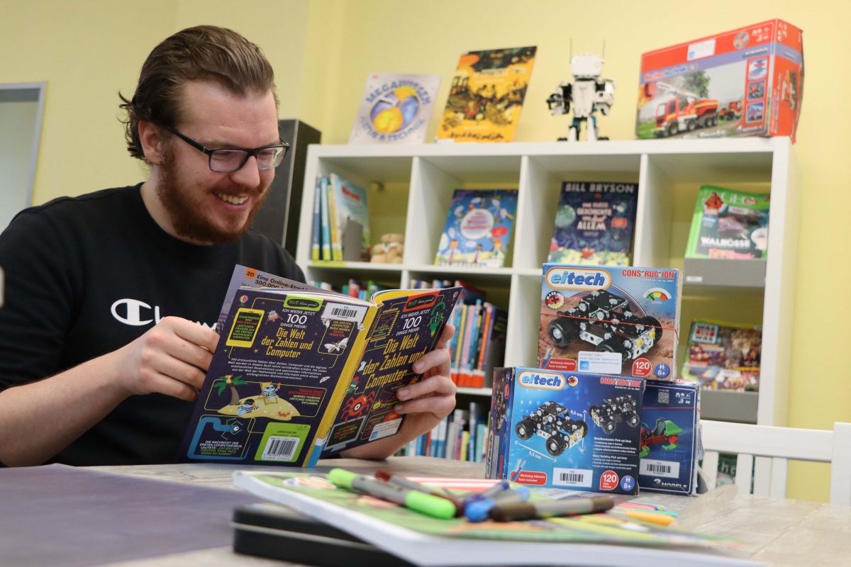 Ein Mann sitzt an einem Tisch und ließt in einem Buch. Im Hintergrund steht ein Regal mit Büchern.