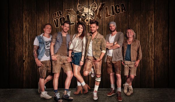 Eine Volksmusikgruppe in Lederhosen posiert vor einer Holzwand mit der Aufschrift: Die VolXXLiga.