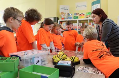 Kinder in orangen T-Shirts und eine Frau stehen an einem Tisch. Sie experimentieren.