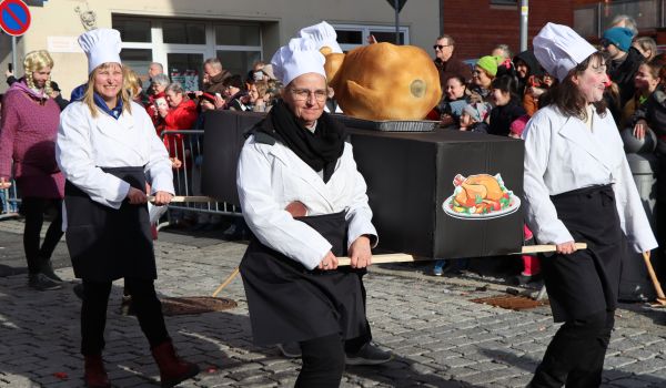 Vier Personen sind als Köche gekleidet. Sie tragen einen großen Gänsebraten.