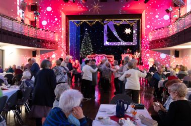 Tanzende Senioren in einem weihnachtlich geschmückten Saal mit Bühne.
