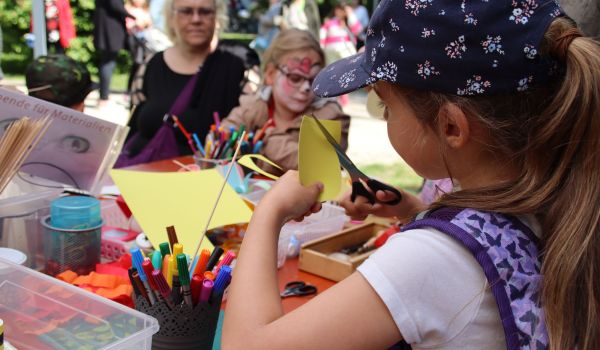 Kinder sitzen an einem Tisch und basteln. Auf dem Tisch sind Stifte, Papier und anderes Bastelzubehör.