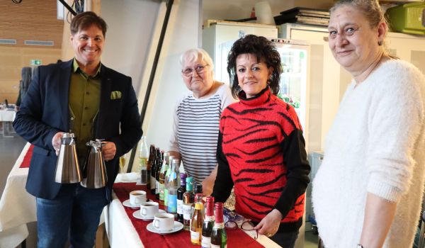 Drei Frauen stehen hinter einer Theke. EIn Mann steht davor und hält zwei Kaffeekannen in den Händen.