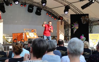 Ein Mann steht auf einer Bühne und spricht in ein Mikrofon. Davor sitzen Menschen.