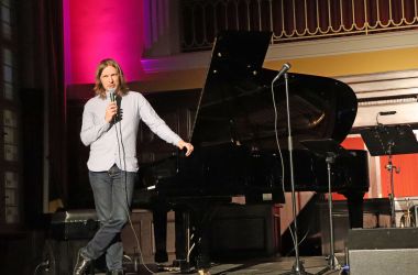 Ein Mann mit einem Mikrofon in der Hand steht auf der Bühne. Er stützt sich mit einer Hand auf einem Flügel ab.