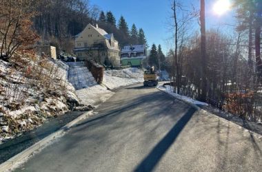 Auf einer Straße steht ein Bagger. Links und rechts von der Straße ist Wald. Es liegt Schnee.