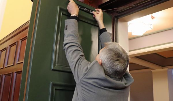 Ein Mann klebt einen Aufkleber mit den Buchstaben und Zahlen: 20 C+M+B 23, an eine grüne Holztür.