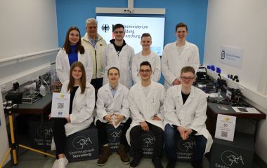 Acht Jugendliche in weißen Kitteln und ein Mann stehen und sitzen nebeneinander.