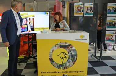 Eine Frau steht hinter einem gelben Messestand. Auf der Standvorderseite ist das Logo der Stadt Sonneberg - das Sonneberger Reiterlein. Daneben steht der Bürgermeister. Im Hintergrund steht ein Bildschirm mit der Aufschrift: Ausbildungsberufe.