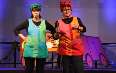 Zwei Frauen stehen auf einer Bühne nebeneinander. Sie sind als Reinigungskräfte verkleidet.