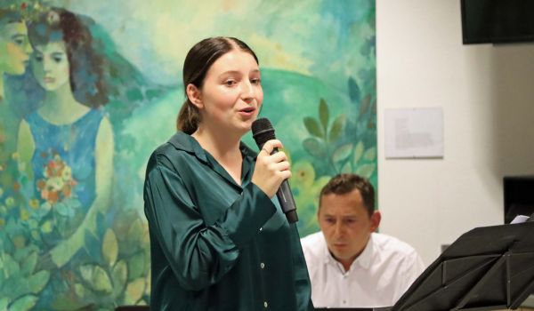 Eine Frau hält ein Mikrofon in der Hand. Hinter ihr sitzt ein Mann am Piano.