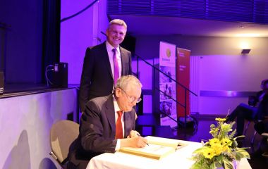 Ein Mann sitzt an einem Tisch und schreibt etwas in ein Buch. Dahinter steht der Bürgermeister der Stadt Sonneberg Dr. Voigt.