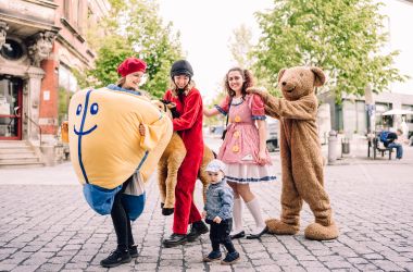 Vier Personen in Spielzeugkostümen stehen in einer Reihe und halten sich an den Schultern. Vor ihnen steht ein kleines Kind.