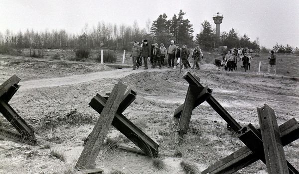Schwarz-weiß: Menschen laufen durch ein ehemaliges Grenzgebiet. Im Hintergrund steht ein Wachturm.