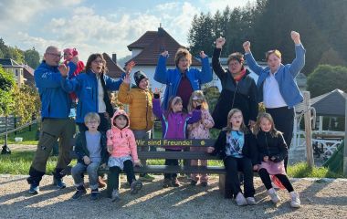 Eine Freunde-Bank wird mit Erwachsenen und Kindern eingeweiht. Sie halten fröhlich die Hände nach oben.