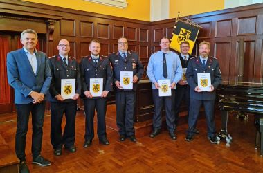 Männer in Feuerwehrunifrom stehen in einer Reihe nebeneinander. Sie halten eine Urkunde in der Hand. Links daneben steht ein Mann im Anzug. Es ist der Bürgermeister der Stadt Sonnberg.