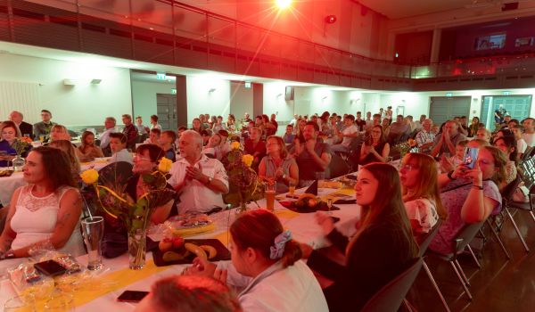 Viele Menschen sitzen an langen Tischreihen in einem großen Saal und klatschen.
