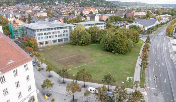 Drohnenaufnahme mit Blick auf das Woolworth-Gelände. Eine Fläche mit Wiese und Bäumen. Dahinter sind Häuser und Gebäude der Stadt zu sehen.