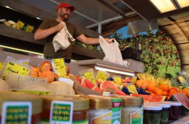 Auf einem Marktstand liegt frisches Obst. Im Hintergrund steht ein Verkäufer.