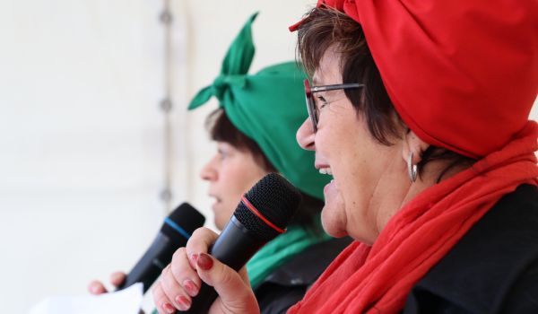 Zwei Frauen stehen auf einer Bühne nebeneinander. Sie sind als Reinigungskräfte verkleidet und halten ein Mikrofon.