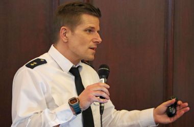 Ein Mann in weißem Hemd steht vor einer Holzwand und hält ein Mikrofon in der Hand