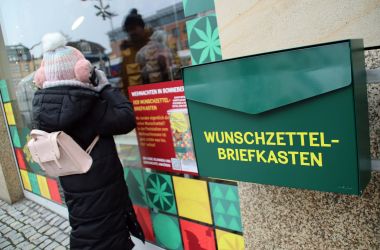 An der Hauswand neben einem Schaufenster hängt ein grüner Briefkasten. Auf dem Briefkasten steht Wunschzettelbriefkasten. Ein Mädchen steht vor dem Schaufenster und sieht hinein.