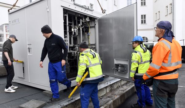 Männer in Warnjacken stehen vor einem Container mit geöffneter Tür. Darin befindet sich eine technische Anlage.