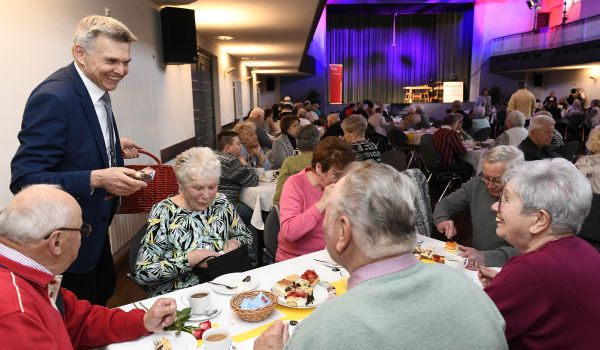 Senioren sitzen an gedeckten Kaffeetafeln in einem großen Saal. Ein Mann verteilt Geschenke. Es ist der Bürgermeister.