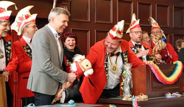 Der Bürgermeister Dr. Heiko Voigt hält ein Steckenpferd in der Hand und lacht. Der Elferrat steht links und rechts daneben.
