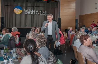 Ein Mann mit Mirkofon ist umgeben von Tischen. An den Tischen sitzen Frauen.