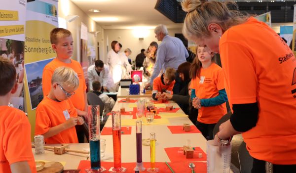 Kinder in orangen T-Shirts machen Experimente an einem Stand.