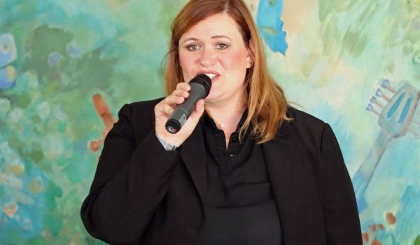 Eine Frau hält ein Mikrofon vor ihrem Mund. Es ist Ines Ehrlicher. Hinter ihr ist eine bunt bemalte Wand.