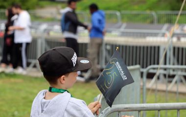 Ein Junge steht vor einer Metallabsperrung und hält ein Fähnchen in der Hand.