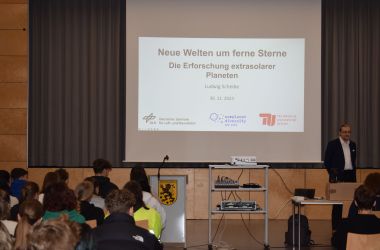 Auf einer Präsentationswand steht: Neue Welten um ferne Sterne. Davor sitzt ein Publikum.