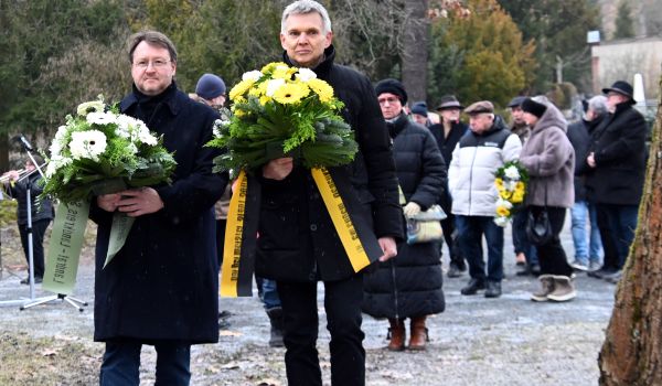 Zwei dunkel gekleidete Männer tragen Blumengebinde zu einem Gedenkstein.