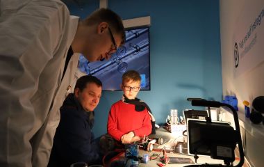 Ein Jugendlicher in weißem Kittel erklärt Experimente an Interessierte.