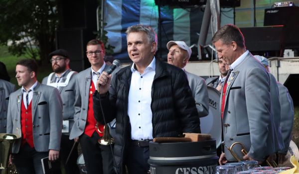 Ein Mann im Anzug hält ein Mikrofon in Hand und steht vor einer Bühne. Es ist der Bürgermeister Dr. Voigt. Hinter ihm stehen Musikanten in Tracht.