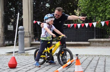 Ein Mann in Polizeiuniform zeigt einem Kind die Richtung. Das Kind trägt einen Helm und sitzt auf einem Fahrrad.