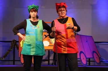 Zwei Frauen stehen auf einer Bühne nebeneinander. Sie sind als Reinigungskräfte verkleidet.