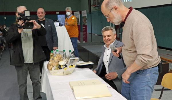 Ein Mann steht mit einem Mikrofon in der Hand hinter einem Tisch. Davor steht ein anderer Mann, der mit einer Kamera filmt.