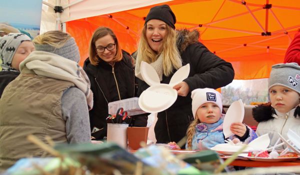 Vor einem Faltpavillon sind zwei Frauen zusehen. Sie basteln zusammen mit Kindern Osterhasen aus Papptellern.