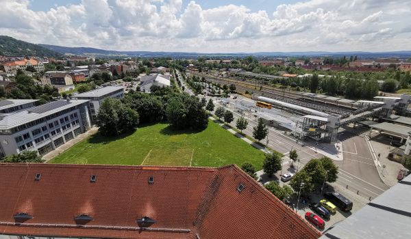 Drohnenaufnahme mit Blick auf das Woolworth-Gelände. Eine Fläche mit Wiese und Bäumen. Dahinter sind Häuser und Gebäude der Stadt zu sehen.