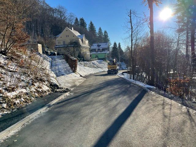 Auf einer Straße steht ein Bagger. Links und rechts von der Straße ist Wald. Es liegt Schnee.