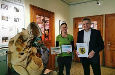 Eine Frau und ein Mann halten eine Urkunde und Mint-Standort-Schilder in den Händen. Der Mann ist der Bürgermeister.