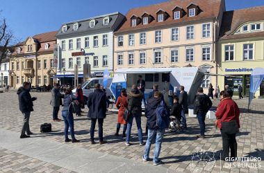 Eine Traube von Menschen steht auf einem Marktplatz vor einem geöffneten mobilen Wagen mit Informationen zum Thema Internet.