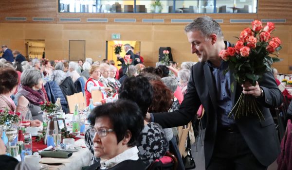 Frauen sitzen an gedeckten Tischen. Ein Mann verteilt Rosen.